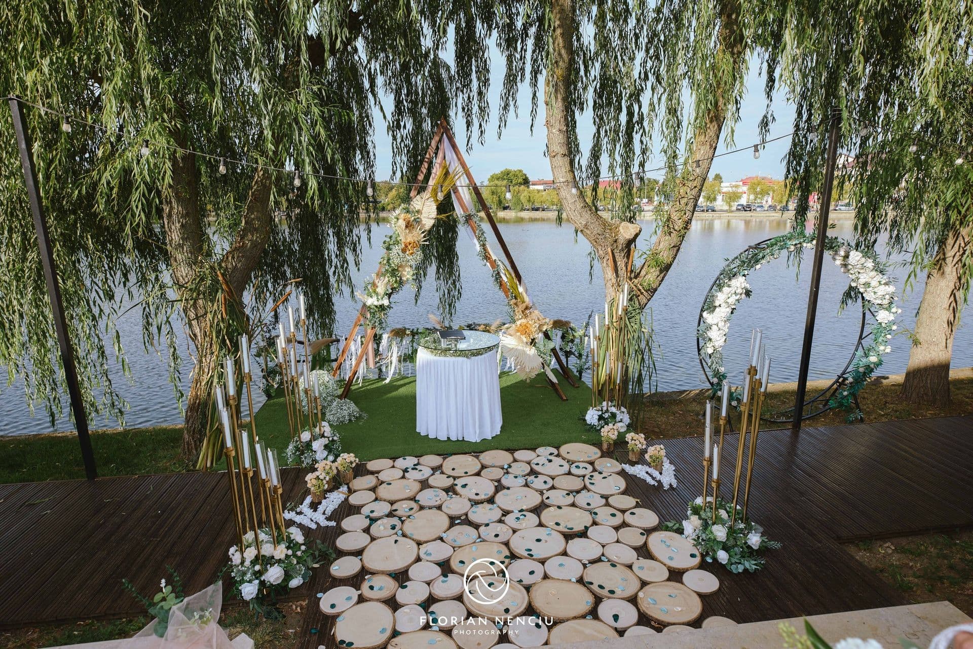 Decor de ceremonie la lac: arcadă triunghiulară din lemn cu flori, potecă din felii de lemn și sălcii
