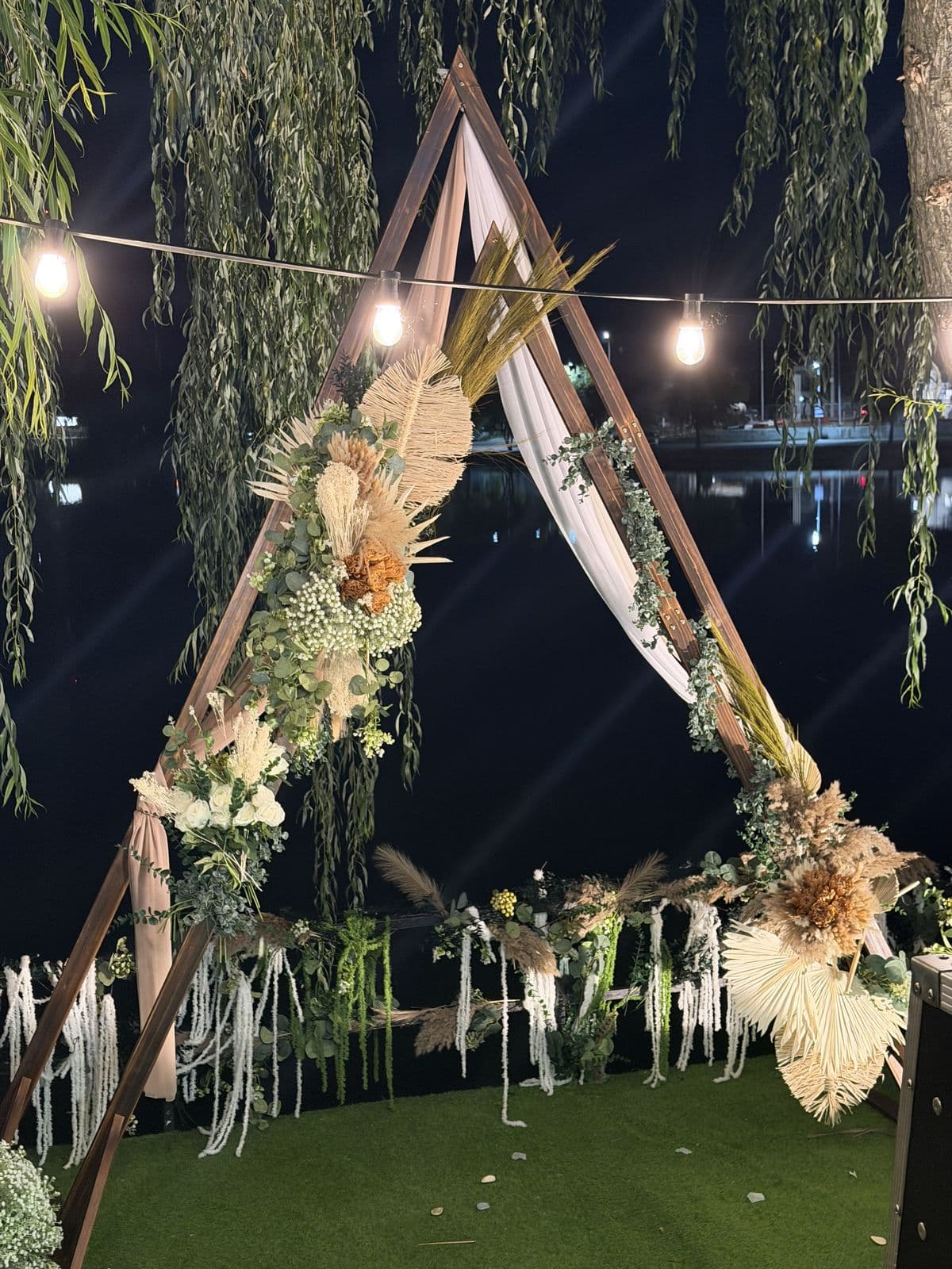 Arcada de ceremonie seara, la malul lacului: lemn, flori uscate și voal, luminată de șiraguri de becuri calde între sălcii.